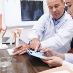 Two medical professionals discussing patient records on a clipboard, with anatomical joint models nearby.