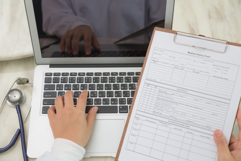 Doctor typing on a laptop while holding a blank Medical History Form on a clipboard.