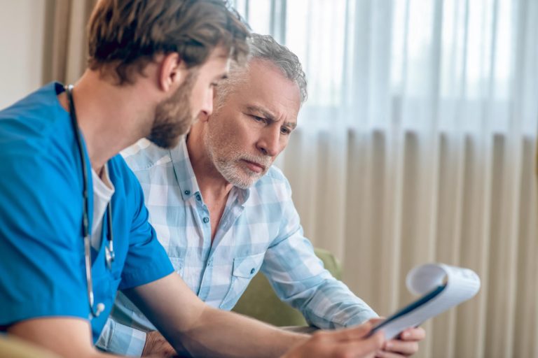 Healthcare professional in blue scrubs reviewing medical documents on a clipboard with a senior man.