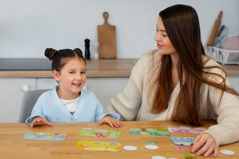 A young girl and a woman sit at a wooden table playing an educational card game together, both smiling. The background shows a kitchen setting with a cutting board and dishes visible.