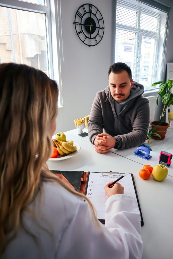 A man sits across a table from a woman in a white coat, likely a nutritionist or hematology doctor, who is writing on a clipboard. Various fruits and a clock on the wall are visible in the bright room.